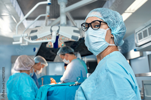 Close-up portrait of a female doctor in glasses and face mask on the background of surgeons team during complex surgical operation in a sterile operating room. Modern medicine, saving patients life.