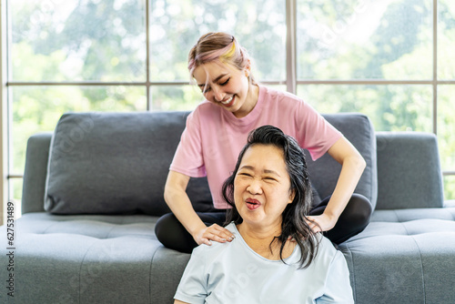 Young Asian daughter massages shoulder, relieves pain of elderly Asian mother while sitting on the sofa at home, Family concept. Health care. Chronic shoulder pain treatment