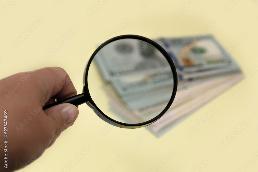 Fototapeta premium hand holding a magnifying glass over a blurred stack of banknotes