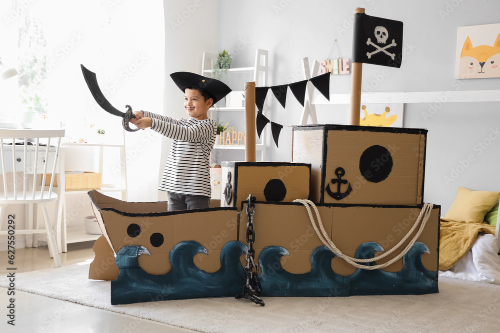 Fototapeta premium Cute little pirate playing with sword in cardboard ship at home