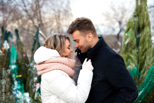 Happy couple in love in Christmas market. Young beautiful man and woman in winterwear embracing and looking at each other with tenderness. Romantic outside dating in winter snowy city.
