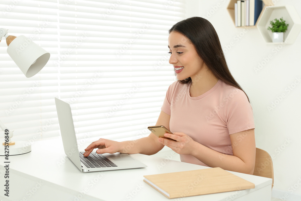 Home workplace. Happy woman with smartphone working on laptop at white desk in room Home workplace. Happy woman with smartphone working on laptop at white desk in room