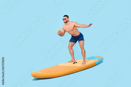 Happy man with refreshing drink balancing on SUP board against light blue background