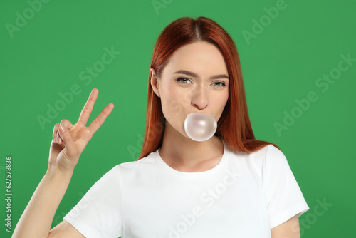 Beautiful woman blowing bubble gum and showing peace gesture on green background