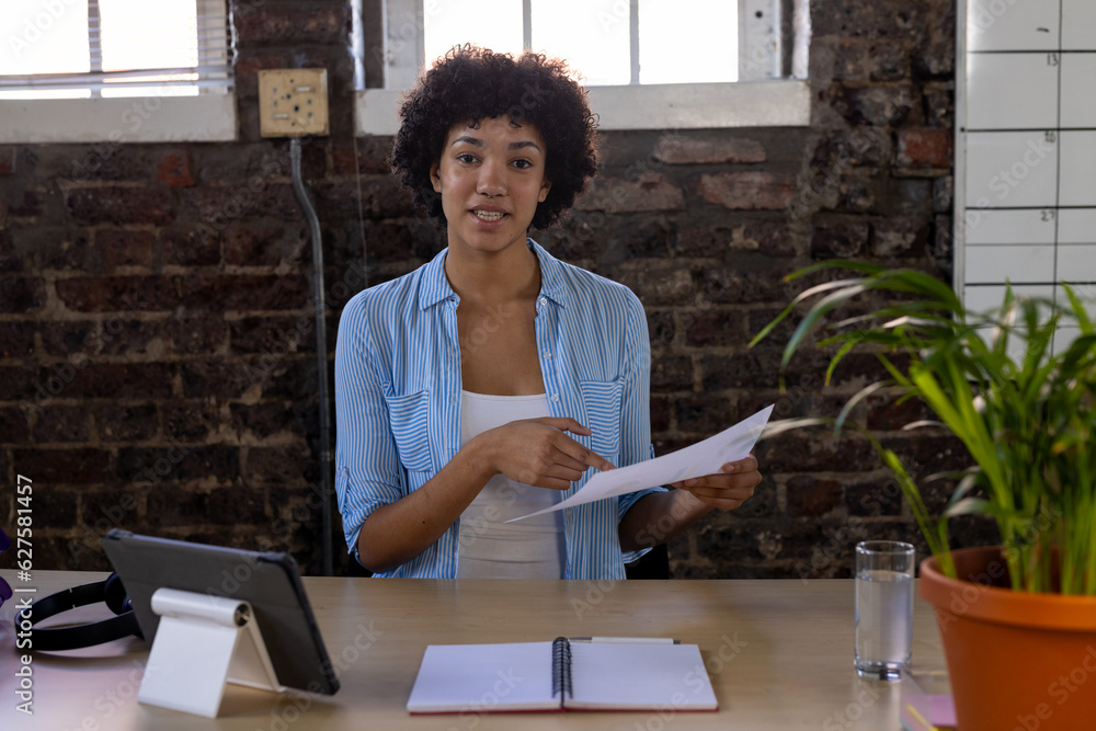 African american businesswoman sitting at desk, having video call and holding documents in office African american businesswoman sitting at desk, having video call and holding documents in office