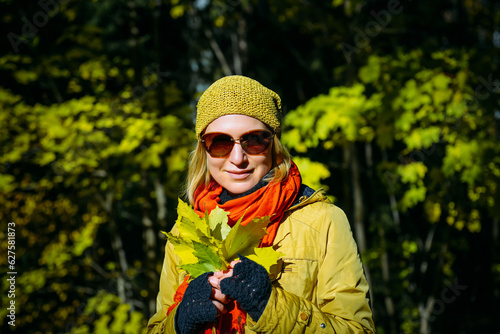 Happy smiling woman wearing bright clothes, holding yellow maple leaves in her hands. Walk in sunny autumn park.