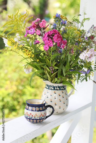 A beautiful ceramic coffee cup and jug with wildflowers on the railing of the summer veranda. Selective focus. 