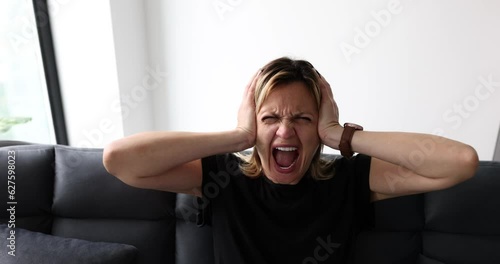 Expressive woman screaming with mouth wide open and covering ears at home on couch. Young woman emotions of anger and hysterics
