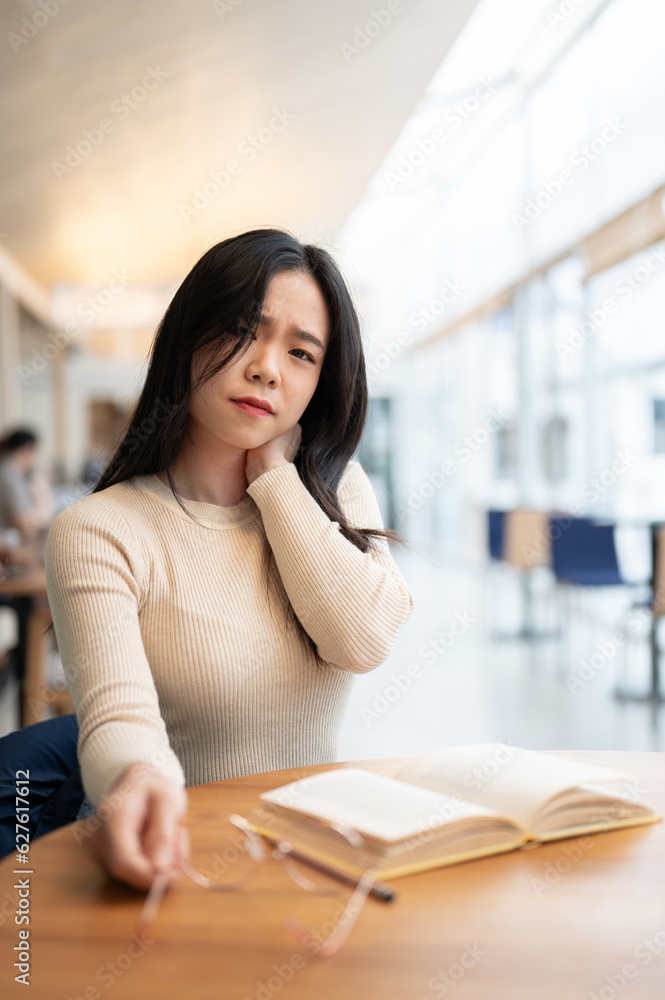 A tired Asian female suffering from a sore neck after a long reading session sitting in a coffee shop A tired Asian female suffering from a sore neck after a long reading session sitting in a coffee shop