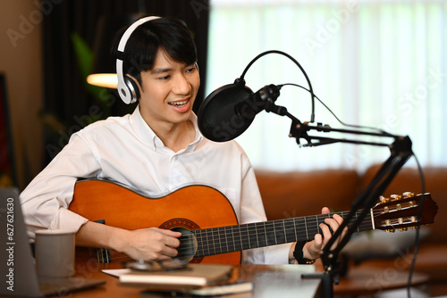 Smiling man in wireless headphones playing guitar during live broadcasting in home studio