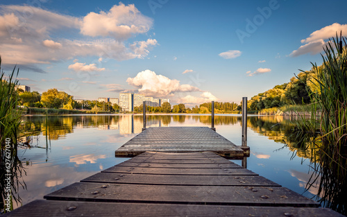 View on Tysiaclecie estate in Katowice, Silesia, Poland. Lightened residential buildings with surrounding trees at afternoon seen through the lake. Little, wooden jetty in the foreground..