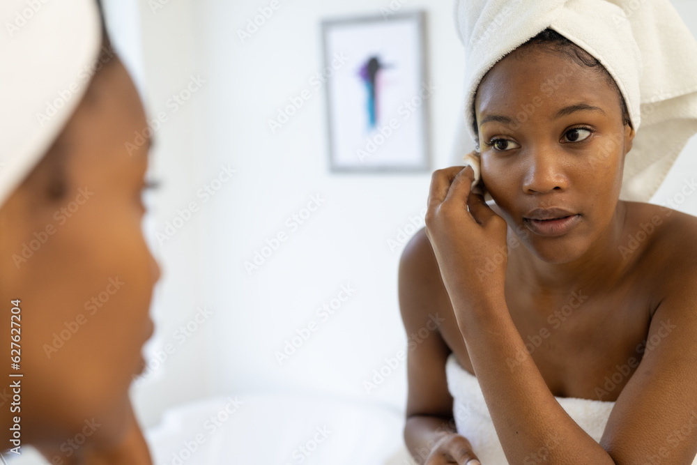 African american woman wearing towel on head cleansing face and looking in mirror in bathroom African american woman wearing towel on head cleansing face and looking in mirror in bathroom