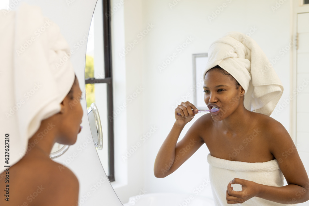 African american woman wearing towel on head brushing teeth and looking in mirror in bathroom African american woman wearing towel on head brushing teeth and looking in mirror in bathroom