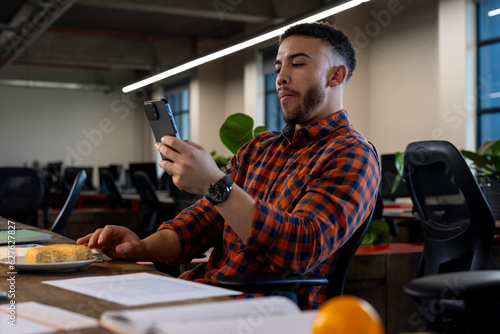 Biracial man using smartphone while having a snack at office photo