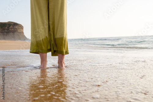 Barefoot white woman steping in the water on the beach on a clear and sunny day. Feet on focus on first view and cliff on the background. She wears green pants and takes place in a sandy beach. © Diego