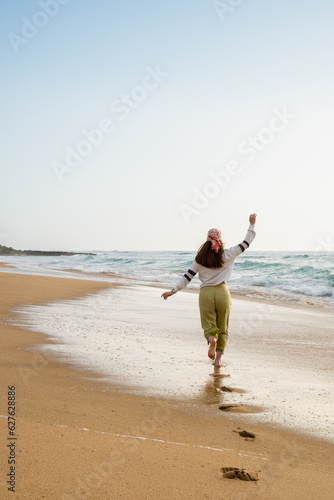 Barefoot white woman running in the beach on a clear and sunny day. Girl on focus on first view and cliff on the background. She wears green pants and white shirt and it takes place in a sandy beach