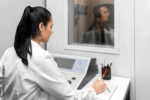 Fotografie, Obraz Audiologist woman doing the hearing exam to a mixed race man patient using an audiometer in a special audio room