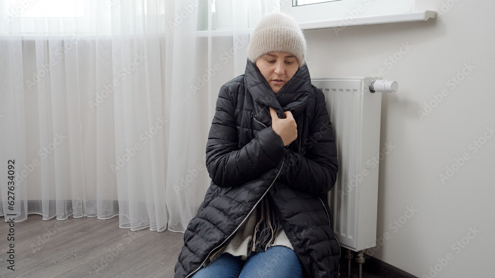 Woman freezing in home putting on warm coat sitting at radiator. Concept of energy crisis, high bills, broken heating system, economy and saving money on monthly utility payments. Woman freezing in home putting on warm coat sitting at radiator. Concept of energy crisis, high bills, broken heating system, economy and saving money on monthly utility payments.