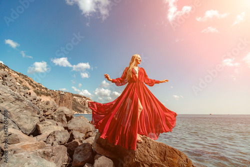 Red dress sea woman. A blonde with flowing hair in a long flowing red dress stands on a rock near the sea. Travel concept, photo session at sea
