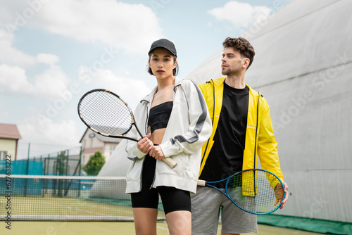 Fototapeta Naklejka Na Ścianę i Meble -  fashionable sportswear, man and woman holding tennis rackets on court,  sport and style