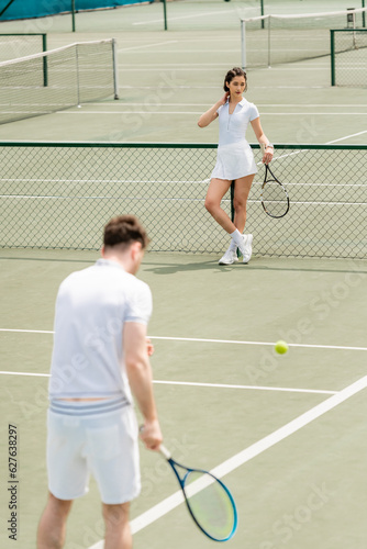 woman standing near tennis net and holding racket, man in active wear on blurred foreground