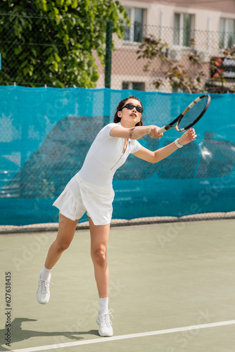 focused woman in sunglasses and active wear holding racket while playing tennis on court, sport