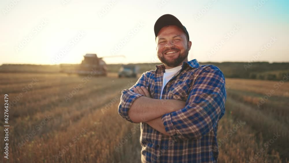 Happy bearded farmer in cap looking at camera standing on agricultural wheat field with combine harvester and dump truck on background. Harvesting, farming at sunset. Food production, agribusiness. Happy bearded farmer in cap looking at camera standing on agricultural wheat field with combine harvester and dump truck on background. Harvesting, farming at sunset. Food production, agribusiness.