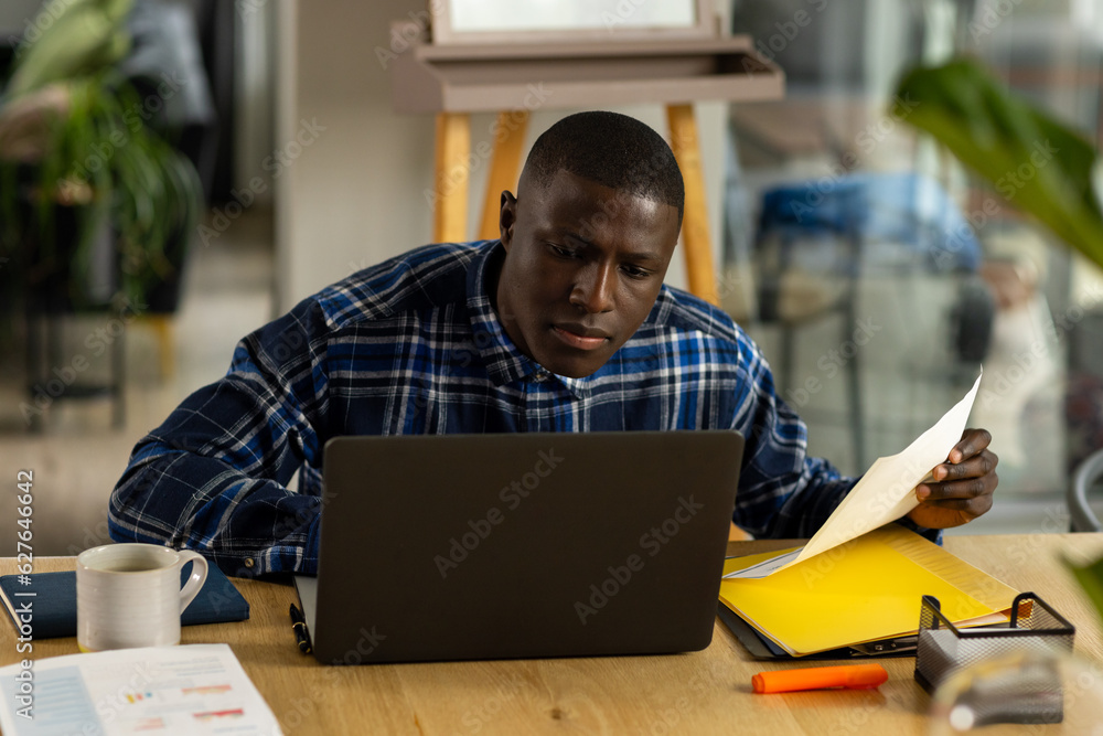 African american man sitting at table using laptop and holding documents at home African american man sitting at table using laptop and holding documents at home