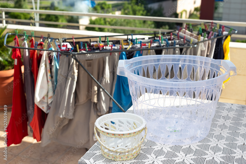 Colorful, clean laundry that has been washed and hung on a metal laundry hanger to dry in the sun on the balcony. Empty laundry basket in selective focus. Routine housework, hygiene concept.