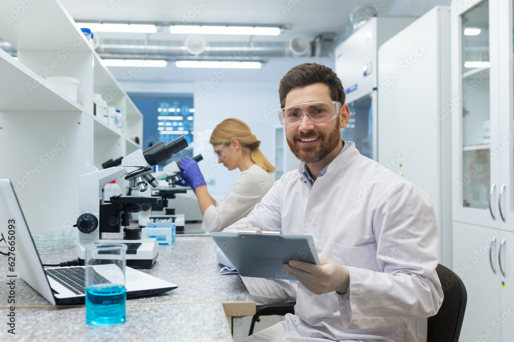Portrait of a young male scientist looking into the camera with a smile. He is sitting in the laboratory in front of a microscope with a documents, a female colleague in the background. Portrait of a young male scientist looking into the camera with a smile. He is sitting in the laboratory in front of a microscope with a documents, a female colleague in the background.
