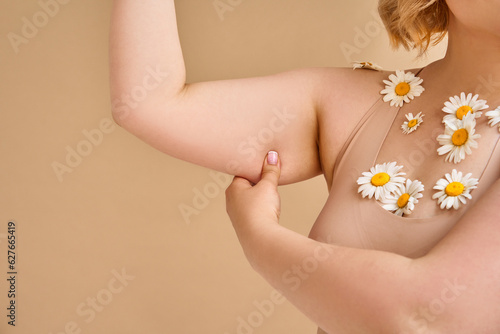 Foto Cropped image of plus size woman showing saggy skin on arm on beige natural background, daisy flowers on chest