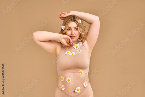 A confident adult blonde woman with beautiful natural body curves poses in comfortable beige underwear with many daisies glued on her body. Women's beauty and body care.Advertising, banner. photo