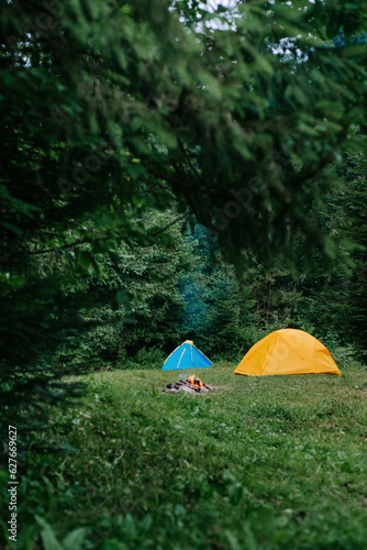 Vertical shot of camp with two tent and warm bonfire near beautiful majestic forest in summertime. Camping lifestyle routine at sunrise. Wanderlust outdoor lifestyle. Camp outdoor mood