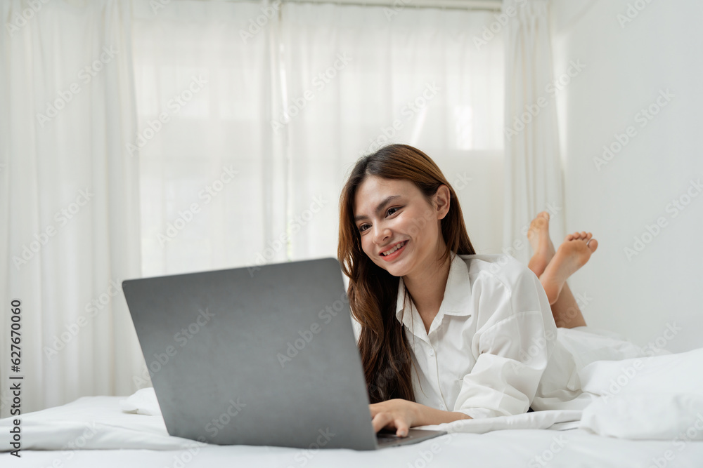 Beautiful happy asian woman lying in bed relaxed and looking at laptop screen. Girl smiling happy while browsing internet on computer, resting at home Beautiful happy asian woman lying in bed relaxed and looking at laptop screen. Girl smiling happy while browsing internet on computer, resting at home