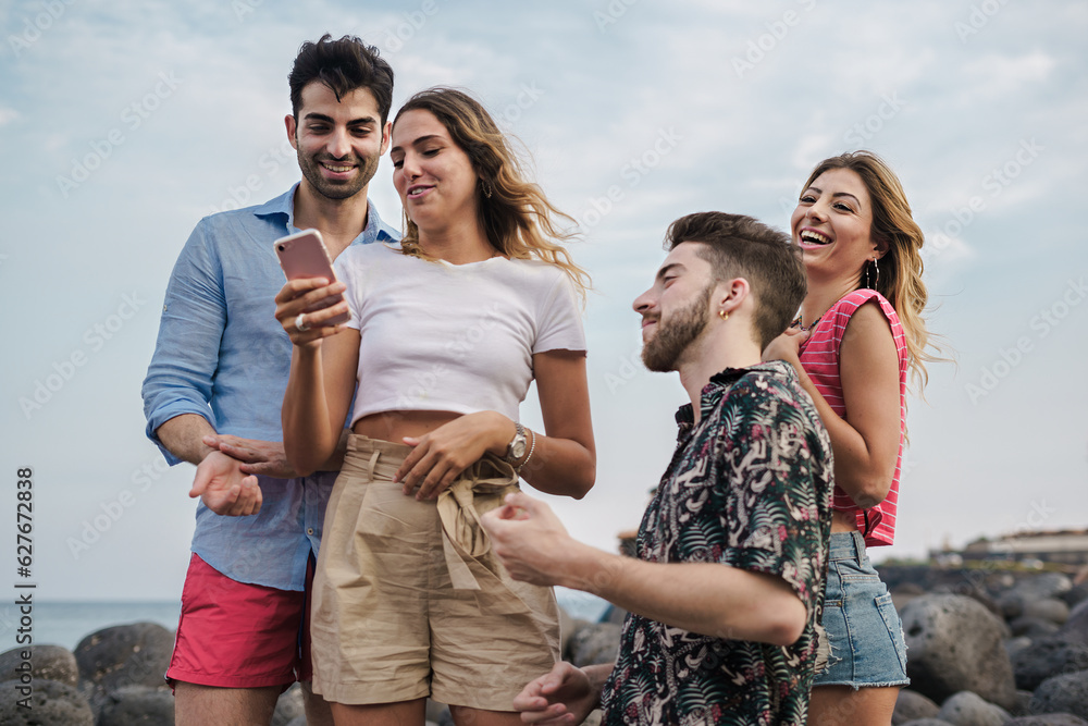 Two Young Couples Having Fun at Seaside with Smartphone - Young Caucasian friends enjoying their vacation at a rocky seaside, laughing and sharing content on a smartphone Two Young Couples Having Fun at Seaside with Smartphone - Young Caucasian friends enjoying their vacation at a rocky seaside, laughing and sharing content on a smartphone