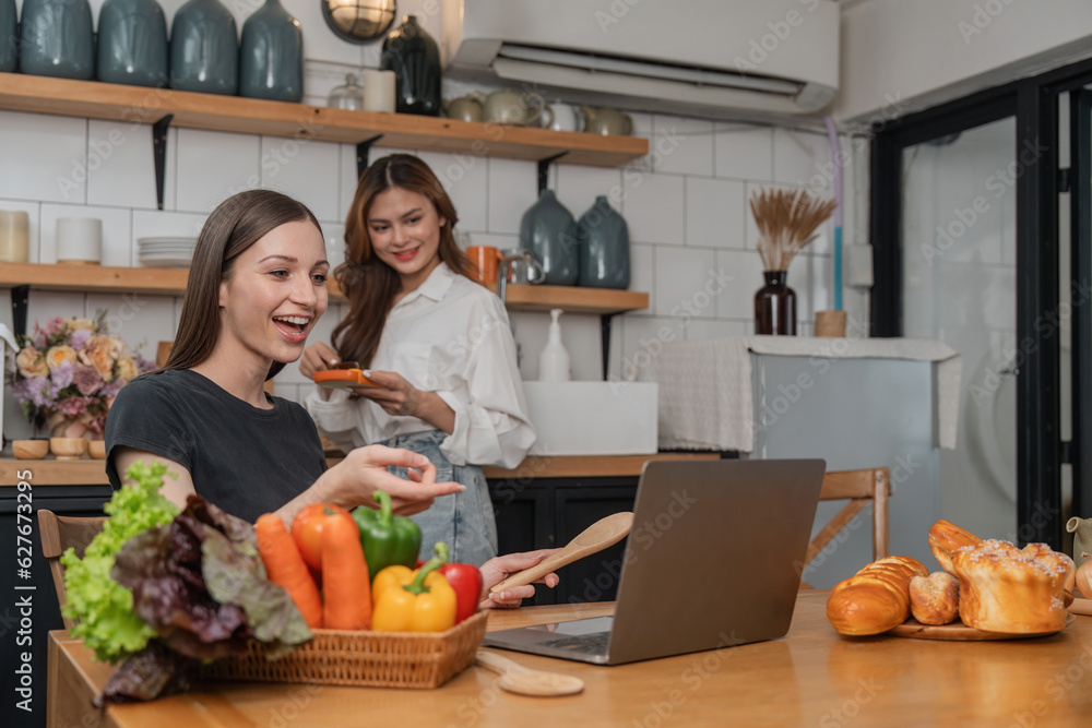 Happy two young women looking laptop computer during cooking together in kitchen room at home. Two young diverse lesbian women spending time together. LGBT and gender identity concept
