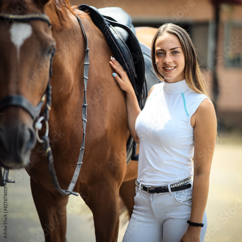 Young rider woman with long hair is standing in the riding stables stroking her horse and smiling at the camera..