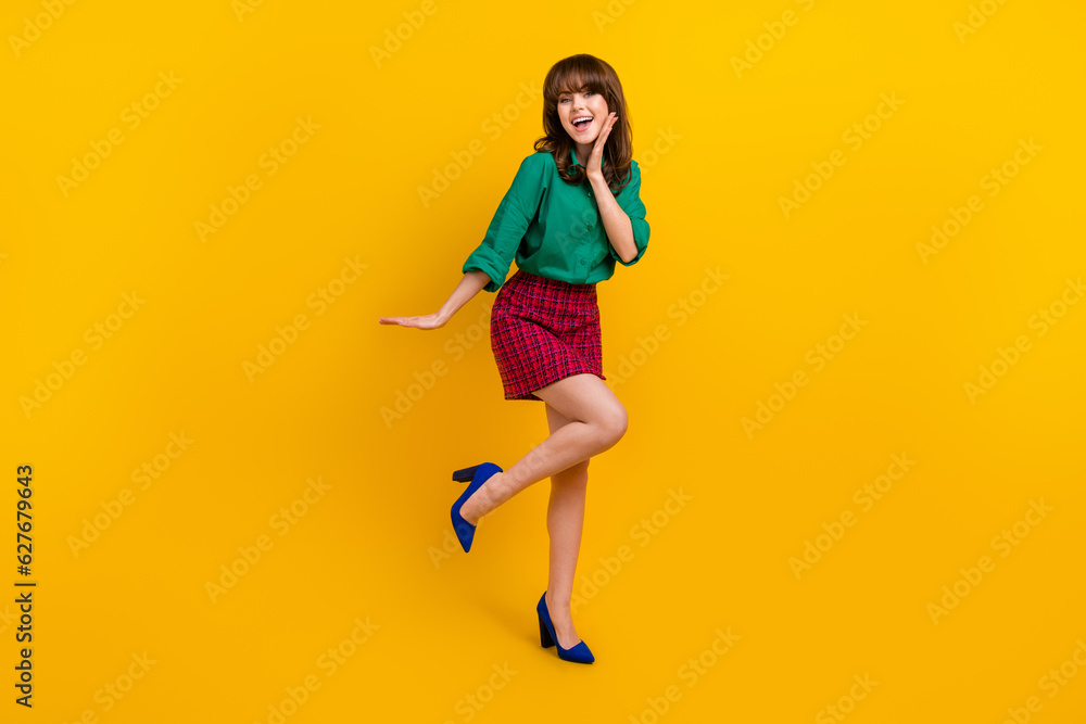 Full size photo of cheerful stunning young girl hand touch cheek stand one leg good mood isolated on yellow color background