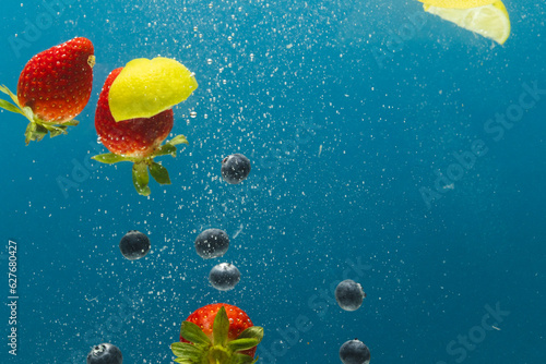 Close up of berries, lime and lemon slices falling into water with copy space on blue background