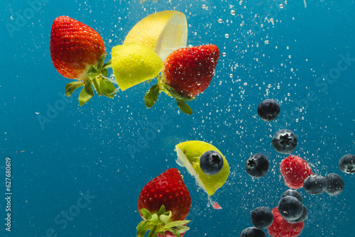 Close up of berries, lime and lemon slices falling into water with copy space on blue background