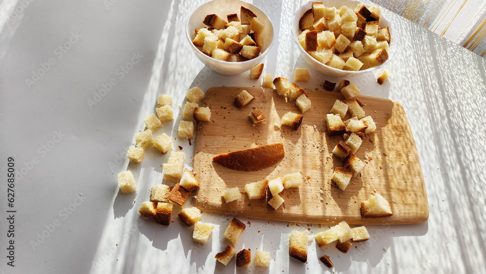 Fototapeta premium Homemade slices of diced dry white rye bread on cutting board and white plate. Crackers for food, healthy eating on table in sunny day, top view