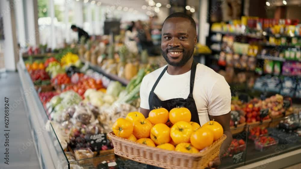 Portrait of a Black-skinned man in a brown T-shirt and black apron posing with a box of yellow tomatoes in his hands in a supermarket Portrait of a Black-skinned man in a brown T-shirt and black apron posing with a box of yellow tomatoes in his hands in a supermarket
