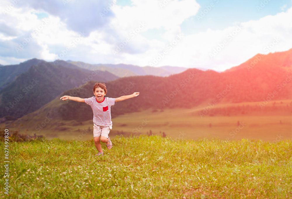Happy little boy running and jumping on green meadow with mountain background