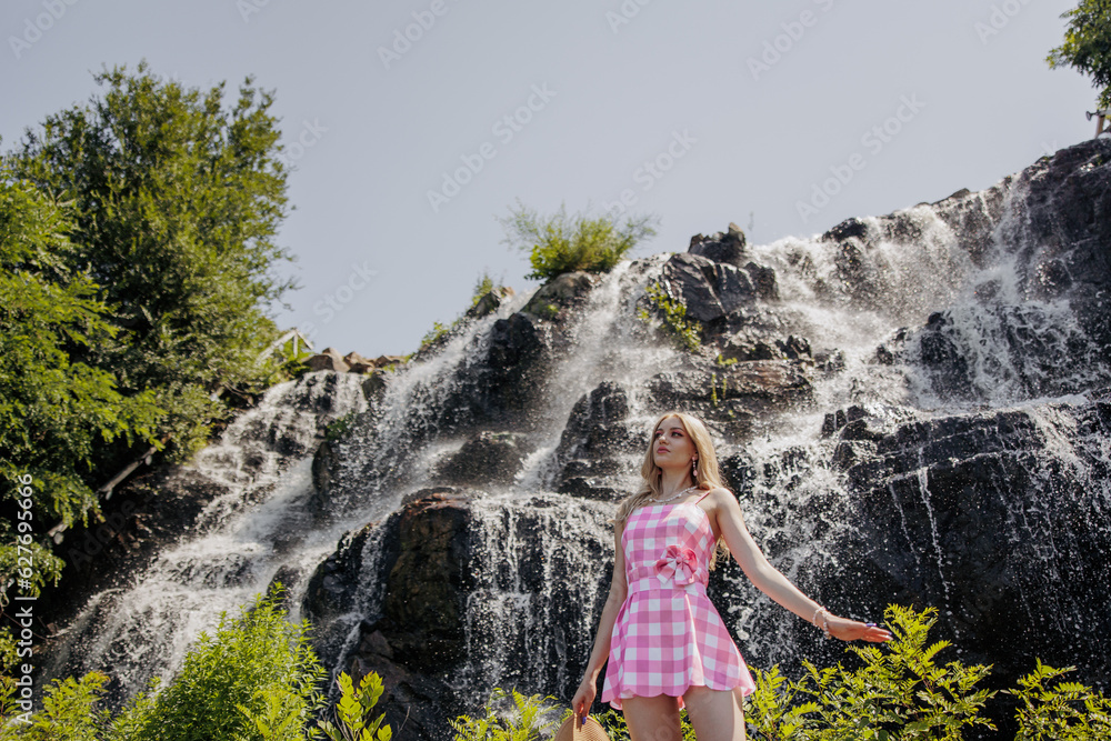 Young blonde girl looking like a Barbie doll in pink mini dress standing against waterfall.