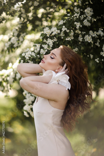 Red-haired girl in a lite dress in blooming apple trees. The girl poses against the background of flowers in the spring park.