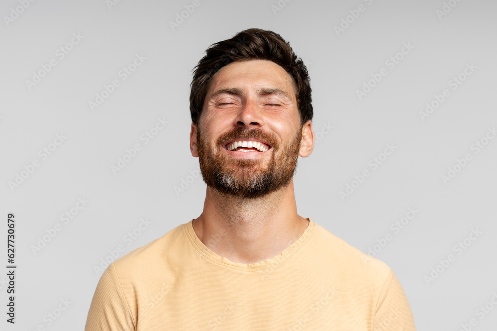 Happy bearded man model posing with closed eyes isolated on gray background. Confident modern hipster with stylish hair enjoying success