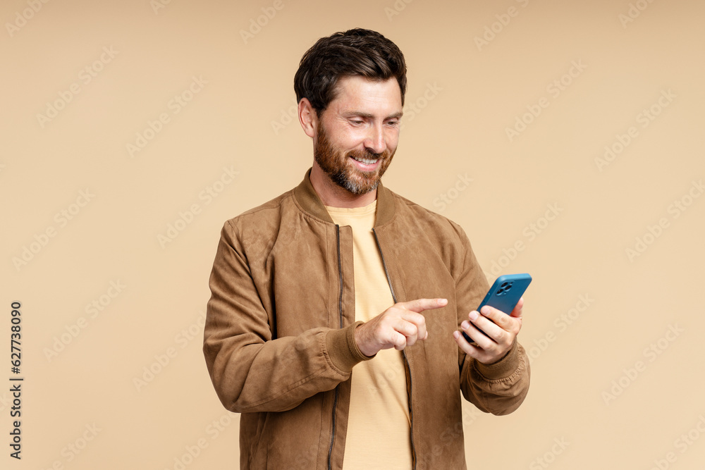 Handsome bearded man in leather jacket holding mobile phone reading text message, communication online isolated on beige background. Hipster guy using mobile app, shopping online, ordering food Handsome bearded man in leather jacket holding mobile phone reading text message, communication online isolated on beige background. Hipster guy using mobile app, shopping online, ordering food