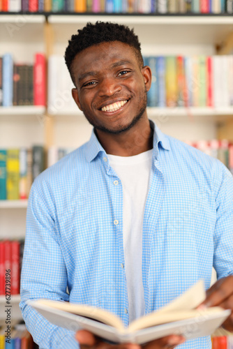Black student in a library holding an open book while wearing a blue collared shirt. The background consists of bookshelves filled with colorful books.