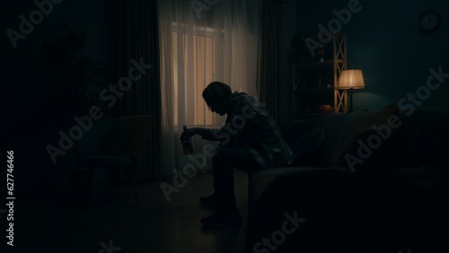 Фотография A devastated young man sits in a room in the dark with a bottle of alcohol