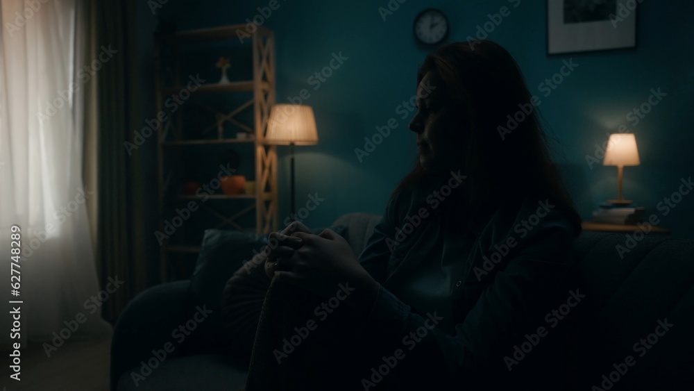 Devastated, detached, lonely young woman sits on a sofa in a dark room close up. Woman desperately thinking about personal problems, going through a crisis. Hopelessness. The concept of mental health.